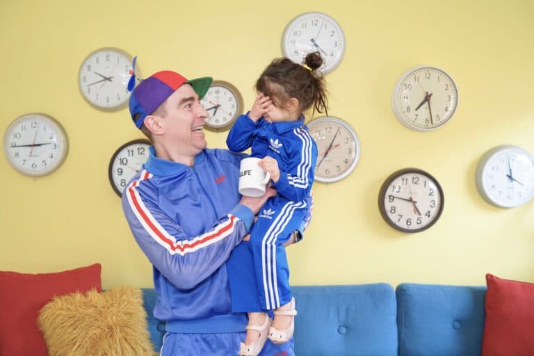 Mann im Sportanzug hält ein Kind - ebenfalls im Sportanzug - im Arm und grinst. Wenn ein Kind auf die Welt kommen, lernen Eltern den Unterschied zwischen früheren Vorstellungen und der Realität.