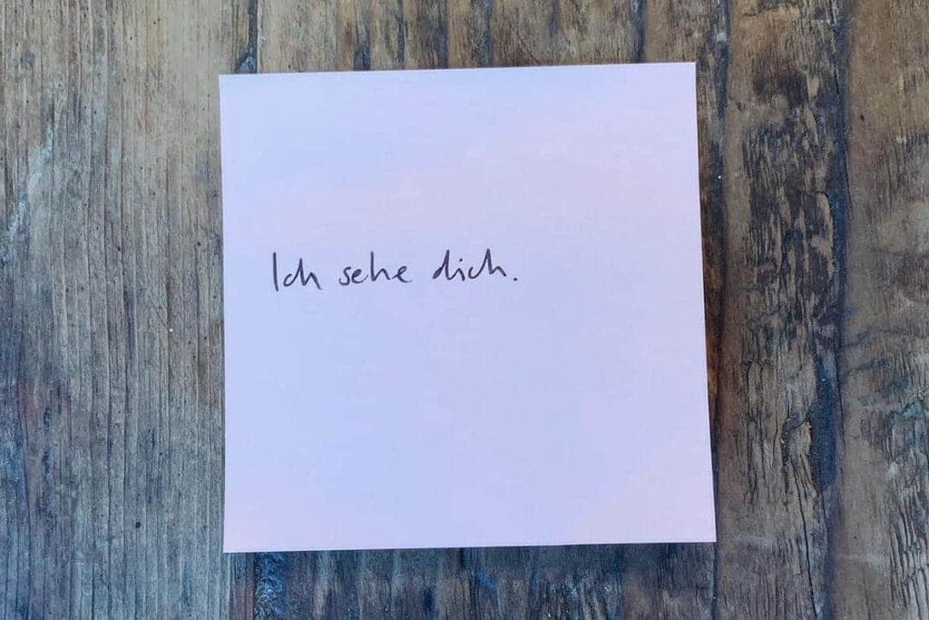 Ein weisser Notizzettel liegt auf einem Holztisch, darauf steht "Ich sehe dich" - eine Liebeserklärung an dich, mich, uns alle