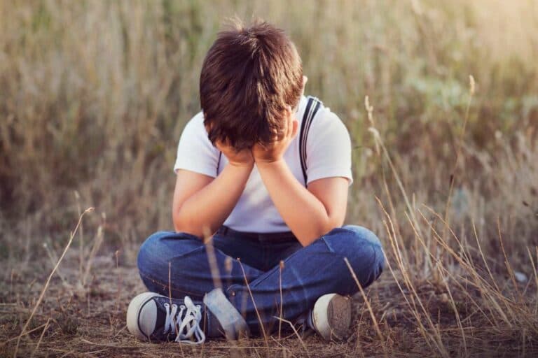 Junge sitzt geknickt in einer Wiese am Boden, sein Gesicht ist in seinen Händen vergraben. – Wie gelingt der Familienausflug? 5 Tipps!