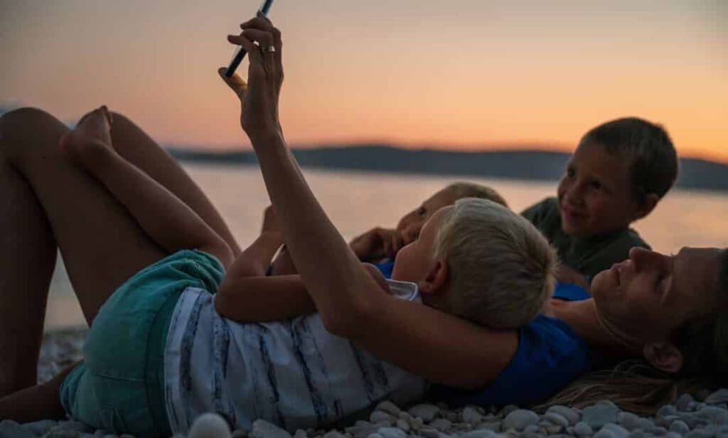 Frau liegt am Boden, mit ihren 3 Kindern im Arm und alle schauen auf ein Smartphone - Tipps für Ferien-Apps