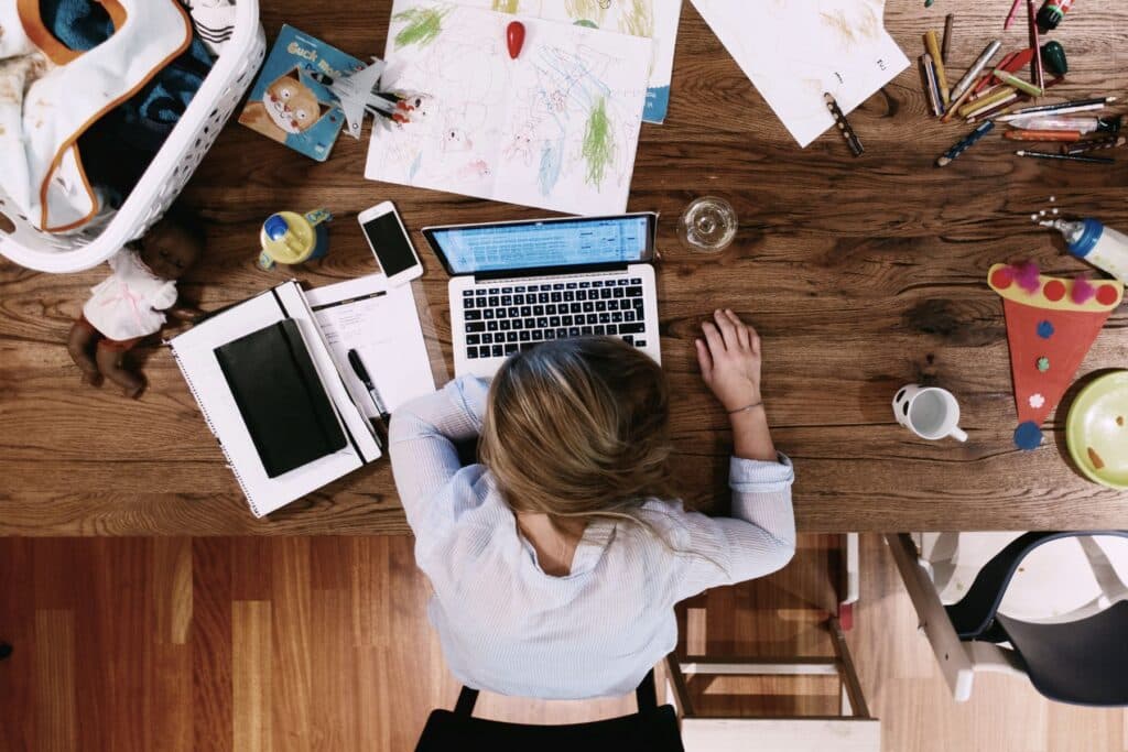 Eine erschöpfte Frau legt ihren Kopf auf den Laptop auf dem Tisch, der übersät ist mit Spielzeug, Schoppen, Wäsche. - Die Working Mom ist ein überholtes Bild.