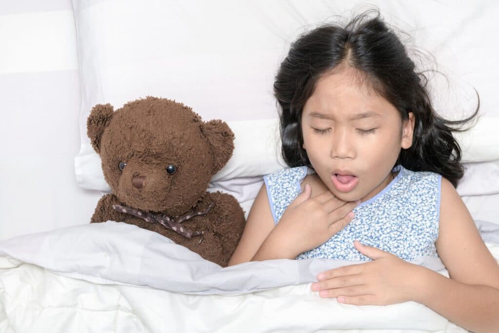 Mädchen liegt im Bett und hat Husten, nebem dem Kind liegt ein Teddybär