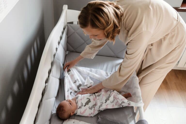 Ein Baby liegt im Bett und wird von seiner Mutter in ein Pucktuch eingewickelt. Ist das Pucken von Babys sinnvoll oder gefährlich?