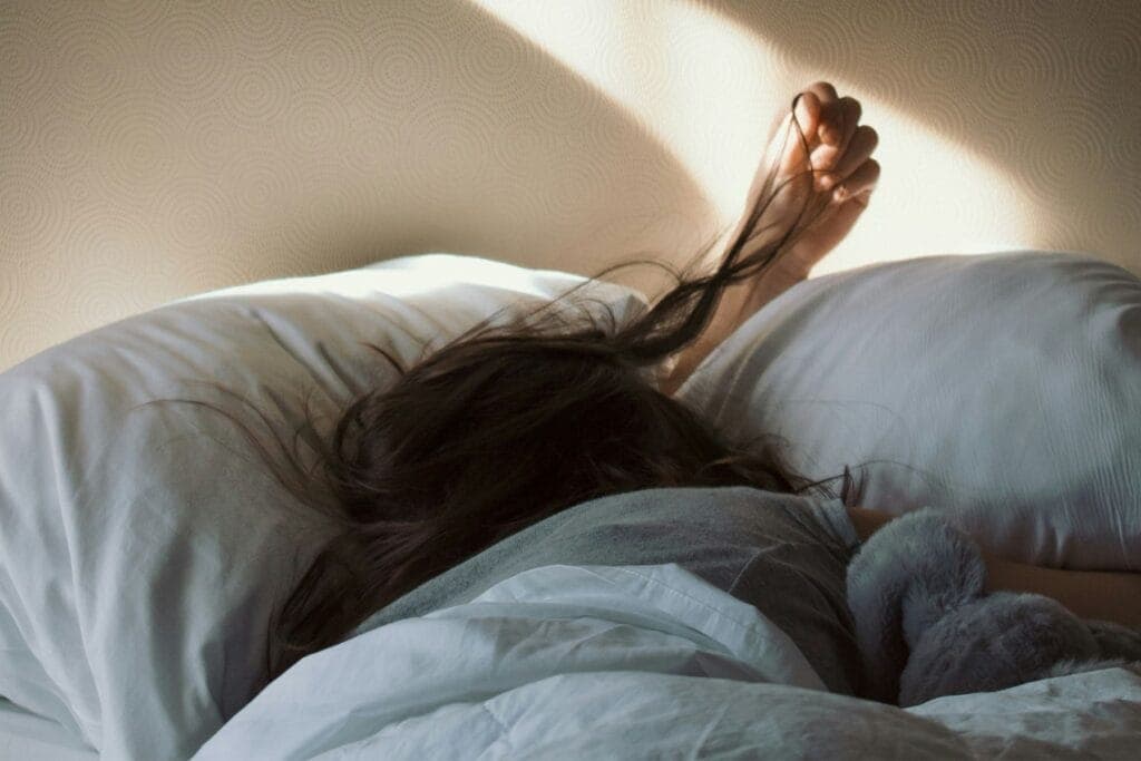 Frau liegt im Bett und hält eine Haarsträhne in der Hand. Schlaf ist wichtig. Trotzdem gehen wir Eltern viel zu oft zu spät ins Bett. Warum?