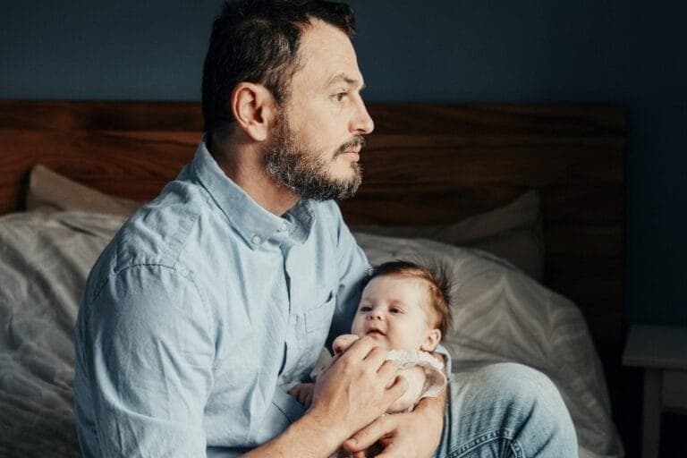 Mann sitzt auf Bett, hält Baby im Arm und schaut nachdenklich in die Ferne. – Alex will ein engagierter Vater sein - und landet im Burn-out.