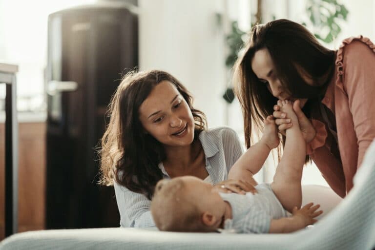 Eine Frau küsst die Füsse eines Babys, eine andere Frau schaut zu. - Wie gelingen Freundschaften zwischen kinderlosen Frauen und Müttern?