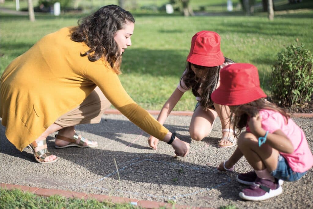 Eine junge Frau und zwei Mädchen malen mit Kreide am Boden. Tipps und Erfahrungen rund um die Anstellung eines Au-pairs in der Schweiz.