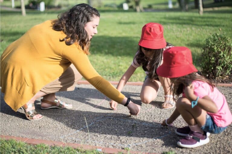 Eine junge Frau und zwei Mädchen malen mit Kreide am Boden. Tipps und Erfahrungen rund um die Anstellung eines Au-pairs in der Schweiz.