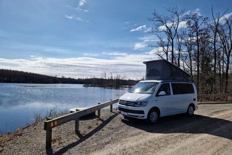 Campervan mit aufgestelltem Dachzelt steht bei Sonnenschein am Rand eines Sees. - Reisen im Camper mit Baby und Kleinkind. Tipps für Eltern.