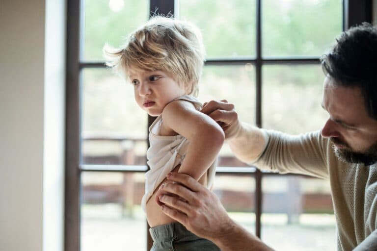 Unglücklich wirkendes Kleinkind kratzt sich am Rücken, Vater untersucht die Stelle. Wie erkennt und behandelt man Neurodermitis bei Kindern?