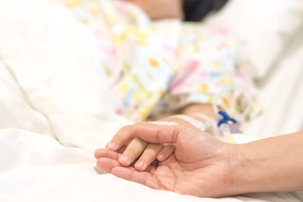Elternteil hält Hand von Kind. Ein Fieberkrampf tritt rasch bei hohem Fieber auf.