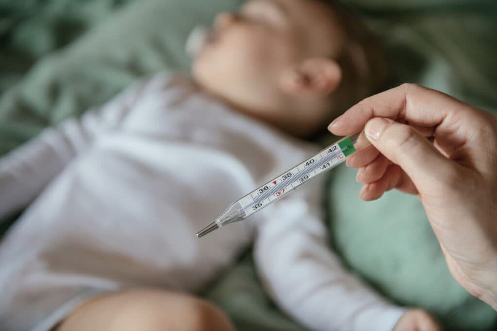 Baby liegt auf dem Bett, im Vordergrund hält eine Hand ein Fieberthermometer. Kinderkrankheiten verunsichern - welche gibt es?