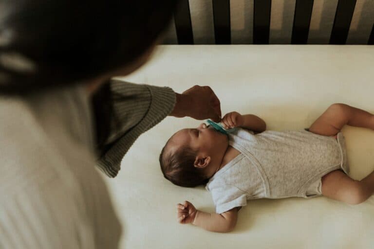 Baby liegt schlafend im Bett, Mutter stabilisiert den Schnuller in seinem Mund. – Die Kinderzahnärztin beantwortet Fragen zum Thema Nuggi.