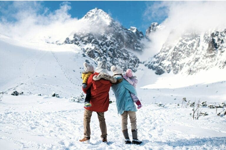 Frau und Mann mit zwei Kindern auf dem Arm stehen in den Bergen im Schnee. Warum die Winterferien uns Eltern gehörig ins Schwitzen bringen.