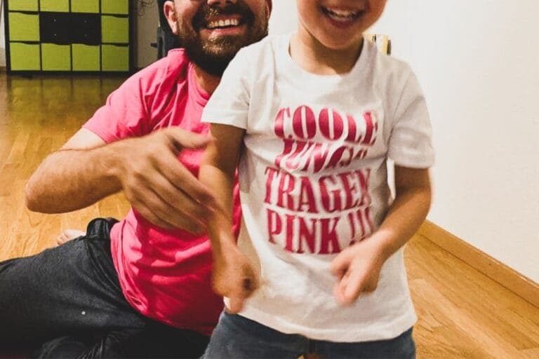 Kleiner Junge und sein Vater, das Gesicht ist abgeschnitten, man sieht nur ihr breites Lachen. Auf dem Kinder-Shirt steht: "Coole Jungs tragen pink!!!"