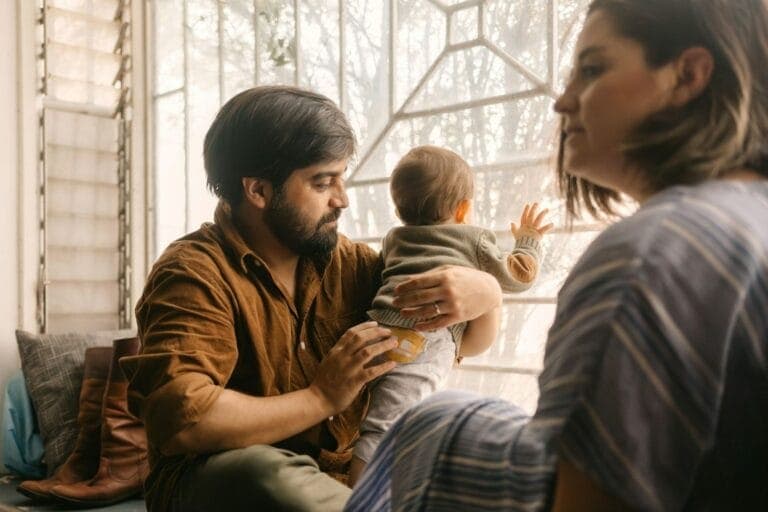 Nachdenkliche Eltern mit Baby sitzen vor einem Fenster – Ist die gleichberechtigte Partnerschaft nur ein Mythos?