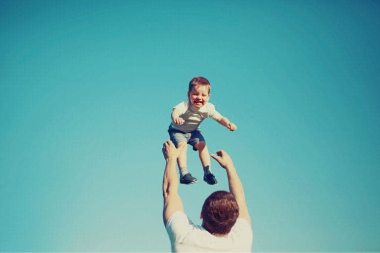 Vater, von hinten sichtbar, wirft seinen lachenden Sohn im Kleinkindalter vor strahlend blauem Himmel in die Luft und fängt ihn wieder auf.