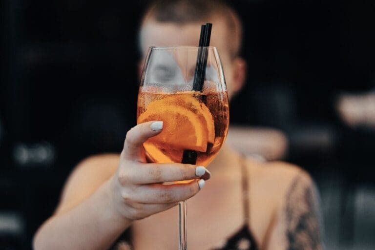 Eine Frau hält ein Glas mit einem orangen Drink in die Kamera. - Alkohol trinken ist ein kontroverses Thema - ist Alkohol Gift oder Genuss?